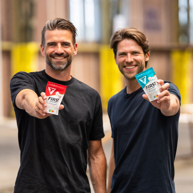 Two men smiling and holding up energy drink packets outdoors.